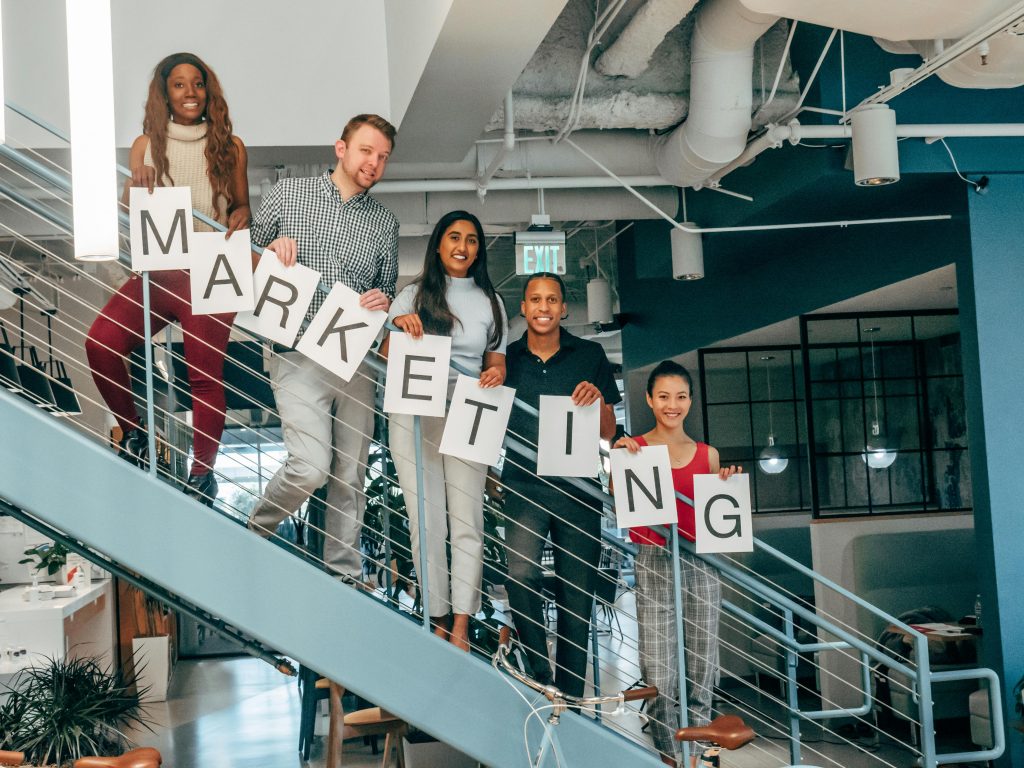 pexels photo 7688468 7688468 Five colleagues smiling and holding a marketing sign on an office staircase, showcasing teamwork.